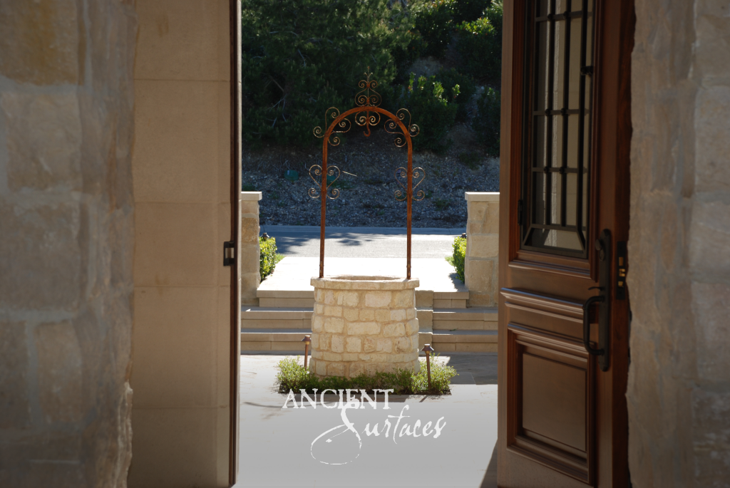 Cylindrical reclaimed limestone well crowned with a wrought iron arch, framed by the open doors of a limestone entry