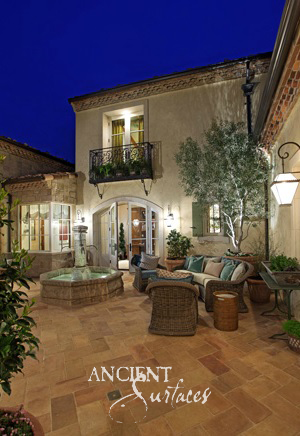 Hexagonal limestone wall fountain lit at night above antique stone paving in a planted courtyard with an olive tree