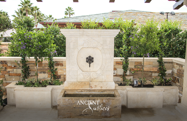 Pale limestone wall fountain centered on a carved roundel above a compact rectilinear basin on a planted upper patio