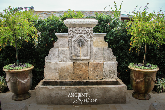 Broad reclaimed limestone wall fountain with a stepped profile, carved cornice and dark iron spout spilling into an aged trough between glazed urns