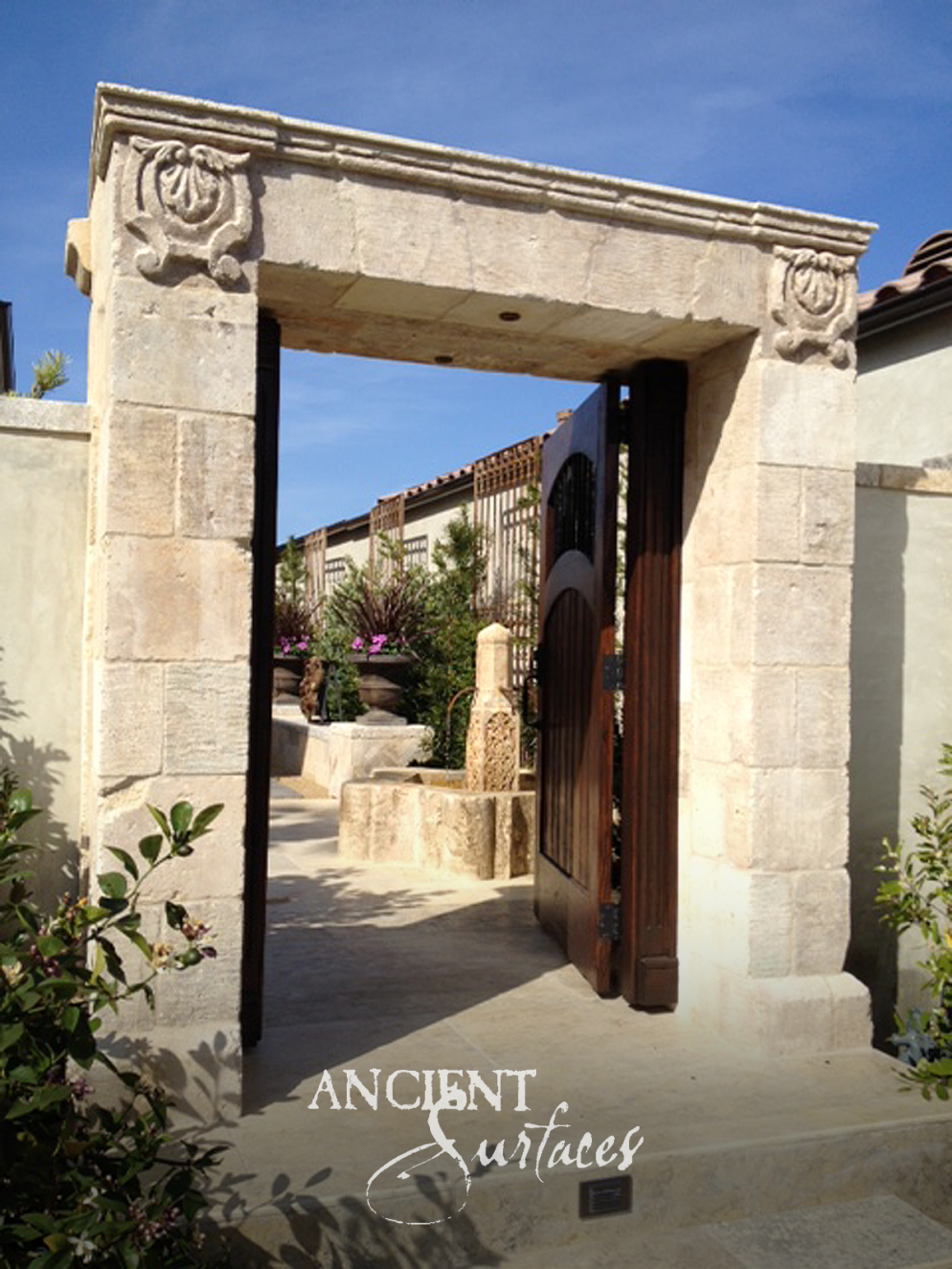 Reclaimed limestone entry portal with a carved lintel and rosette blocks framing a timber gate and a courtyard fountain beyond